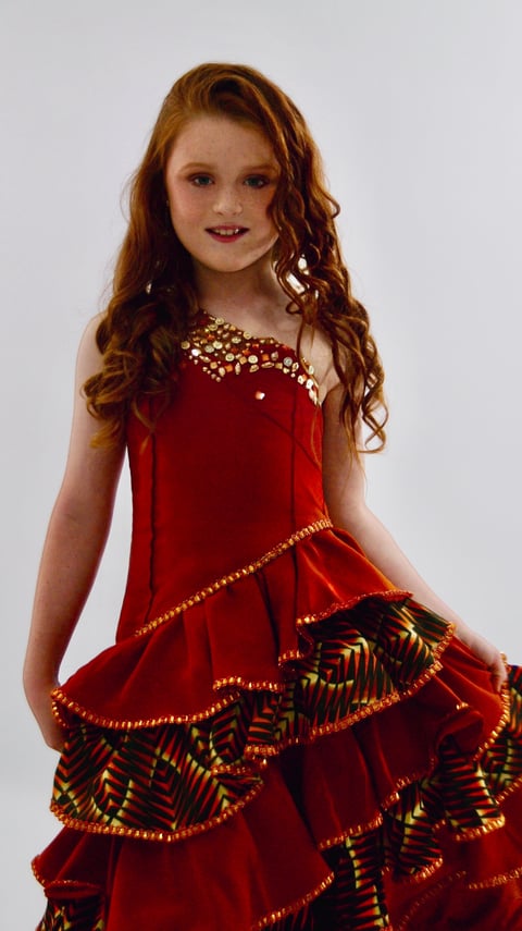 Young girl with long reddish-brown curly hair smiling at camera wearing ornate red dress with gold beaded details