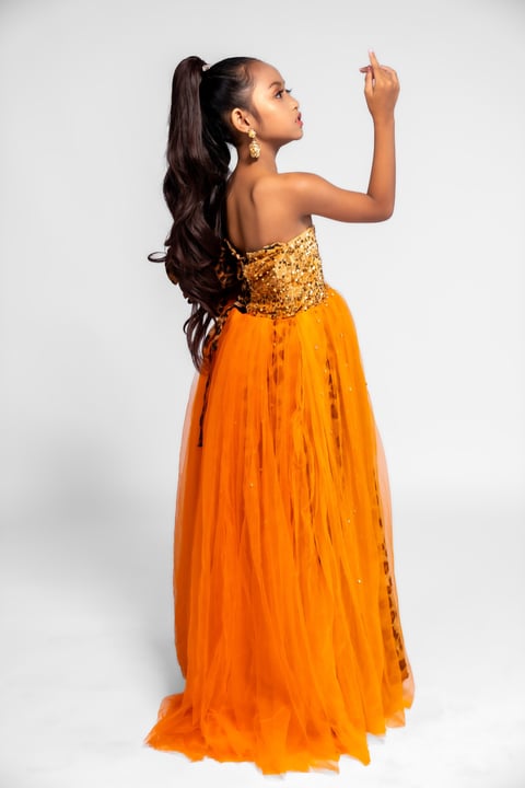 Woman in orange floor-length gown with gold beaded bodice, long dark ponytail, posing against white background