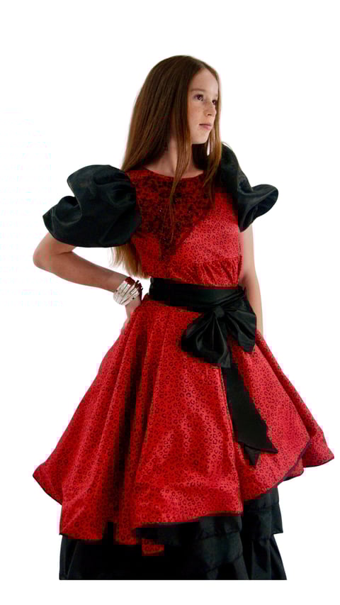 Young woman wearing a red sparkly dress with black off-shoulder ruffles, black bow belt, and holding a black hat, standing against white background