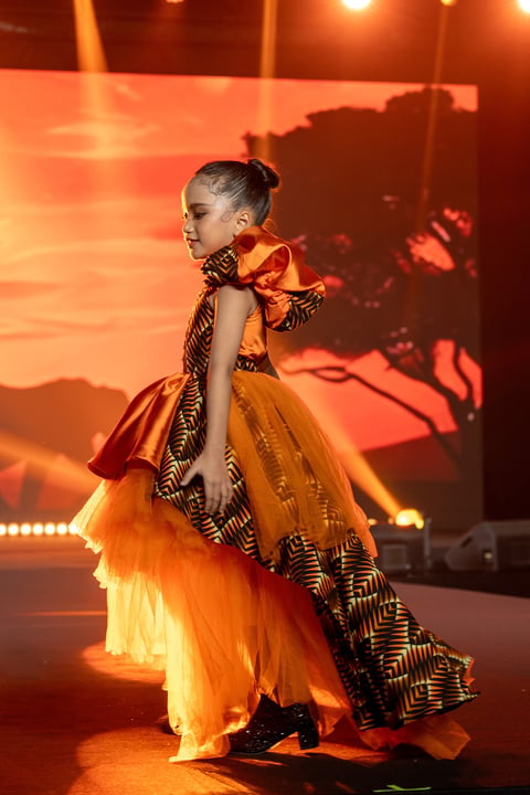Young girl in vibrant yellow tulle dress with orange and striped accents performing on stage under warm orange lighting.