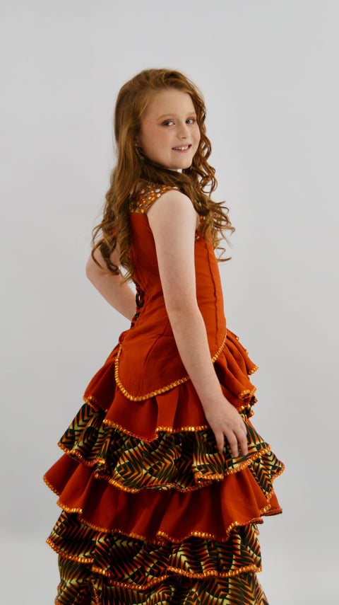Young girl with reddish-brown curly hair wearing a red and gold tiered dress, smiling and looking over her shoulder against a gray background