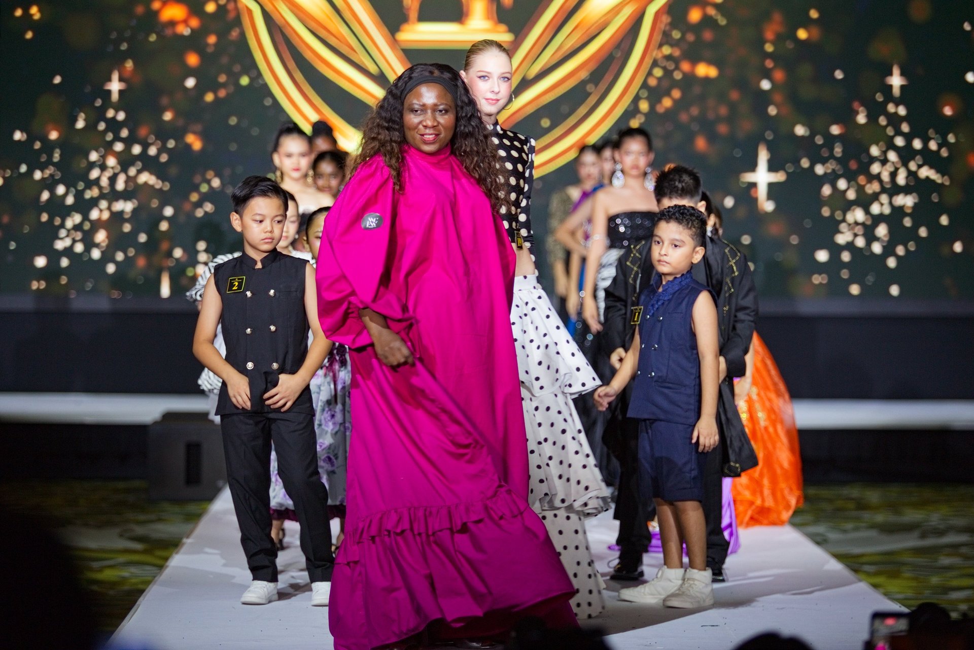 Woman in vibrant magenta gown leading children down a glittering runway during a fashion show with golden stage backdrop