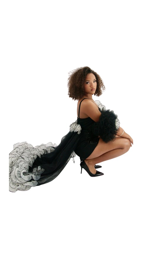Woman with curly brown hair wearing a black dress with tulle details, posing in a crouching position against a white background