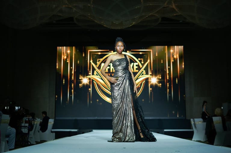 Model walks runway in shimmering gray gown with gold geometric backdrop and dramatic lighting at fashion show