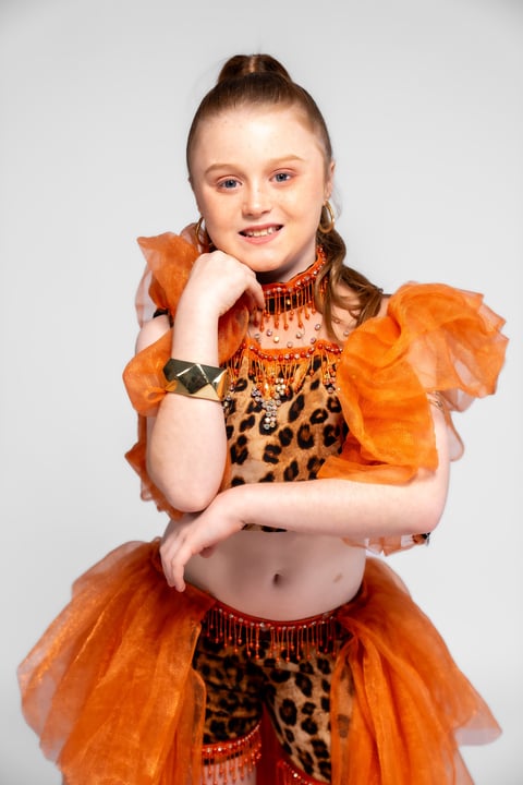 Young girl in orange and leopard print costume with ruffled sleeves, posing with hand on chin against white background