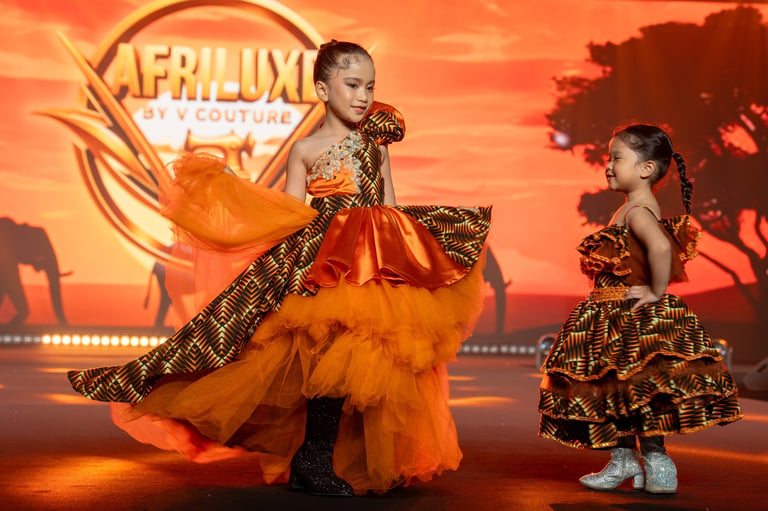 Woman and child in matching orange and gold patterned dresses pose in front of an Afro Luxe Couture backdrop with warm sunset lighting.