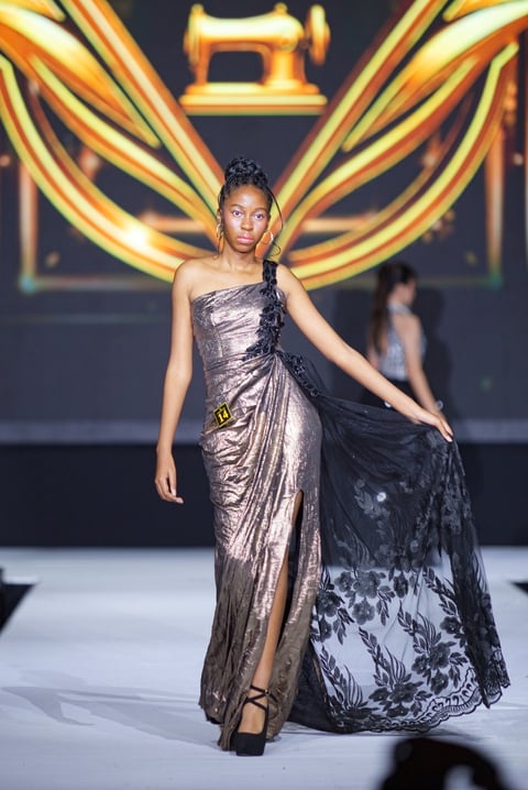 Model on runway wearing metallic silver and black evening gown with dramatic sleeves against glowing gold neon backdrop