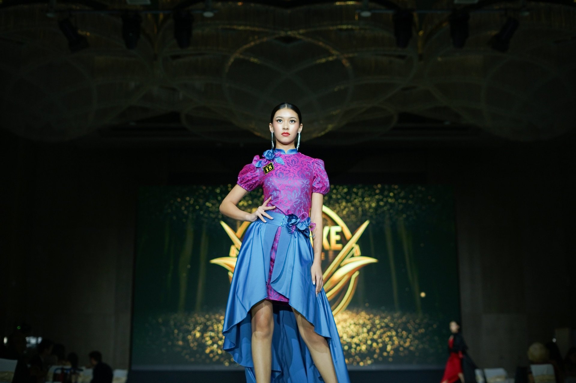 Model on runway wearing pink puffed sleeve top and blue asymmetrical skirt at fashion show with gold backdrop