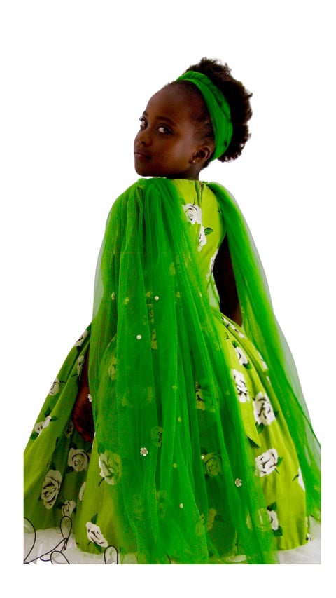 Young girl in green floral dress with matching headband and flowing cape, standing against white background