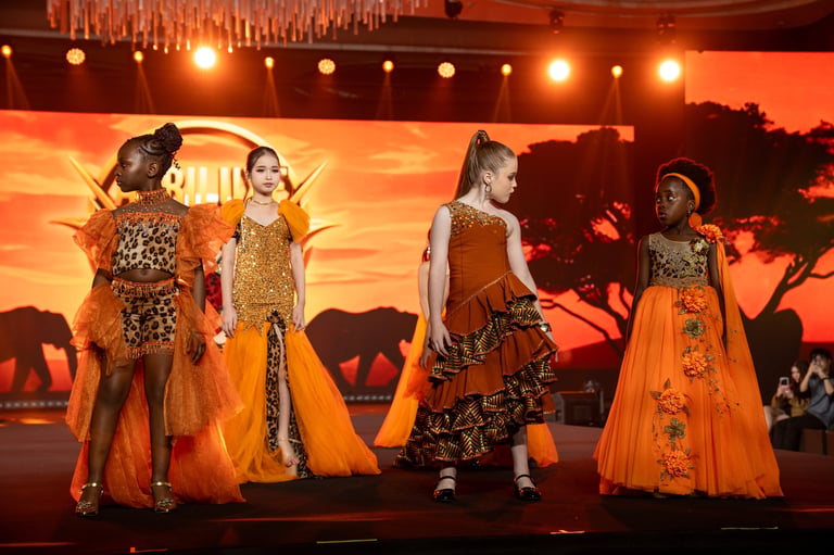 Four young girls wearing ornate orange and gold costumes perform on stage with an African savanna backdrop and sunset scenery.