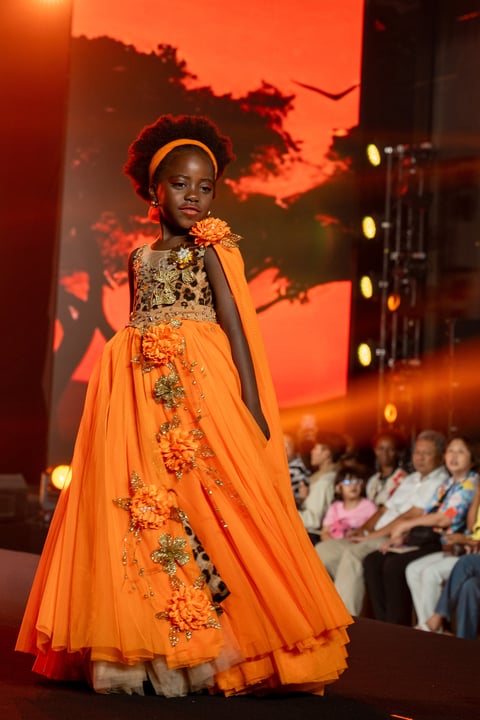 Young girl on fashion runway wearing an elegant orange gown with floral embellishments and a gold headband against a warm-lit backdrop