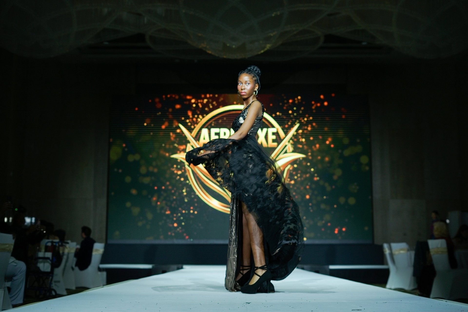 Model on runway wearing black sequined gown with gold accents and Lakers logo backdrop