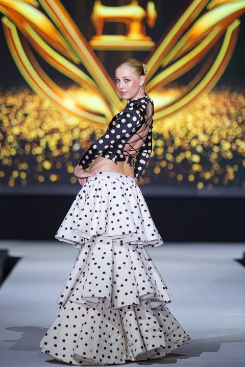 Model on runway wearing black polka-dot crop top and tiered white polka-dot skirt against gold lighting backdrop