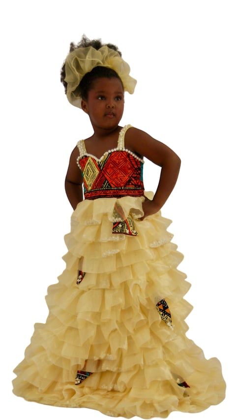 Young girl wearing a formal cream and red dress with ruffled layers, matching headpiece, standing against a white background