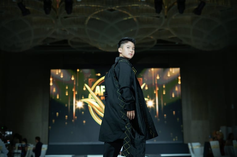 Model wearing black coat on runway during fashion show with illuminated backdrop and audience in background