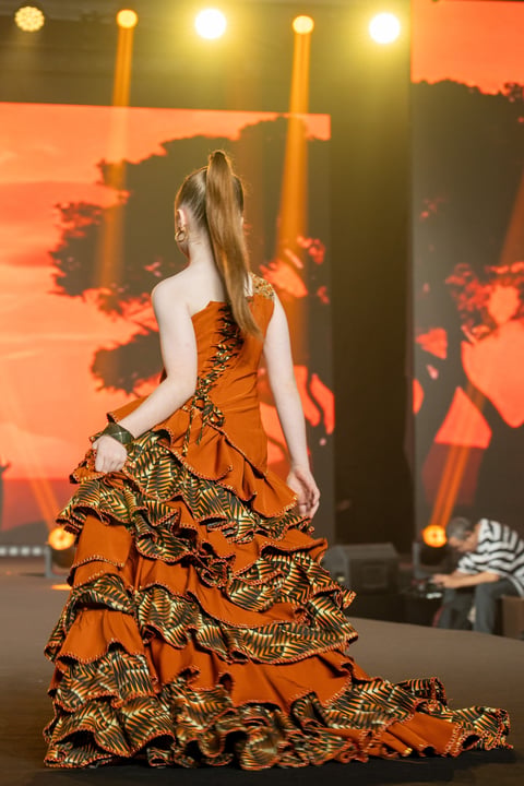 Model wearing orange ball gown with gold patterned ruffles on runway with dramatic stage lighting