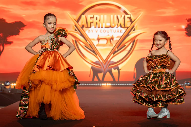 Two young girls in elegant formal dresses standing in front of a golden Afrilux logo against an orange sunset background