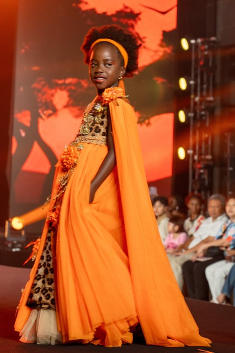 Child model walks runway wearing vibrant orange cape and ornate patterned dress with gold accents under stage lighting