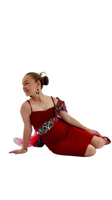 Young woman sitting on white background wearing red dress with decorative belt and matching red ballet pointe shoes, smiling at camera