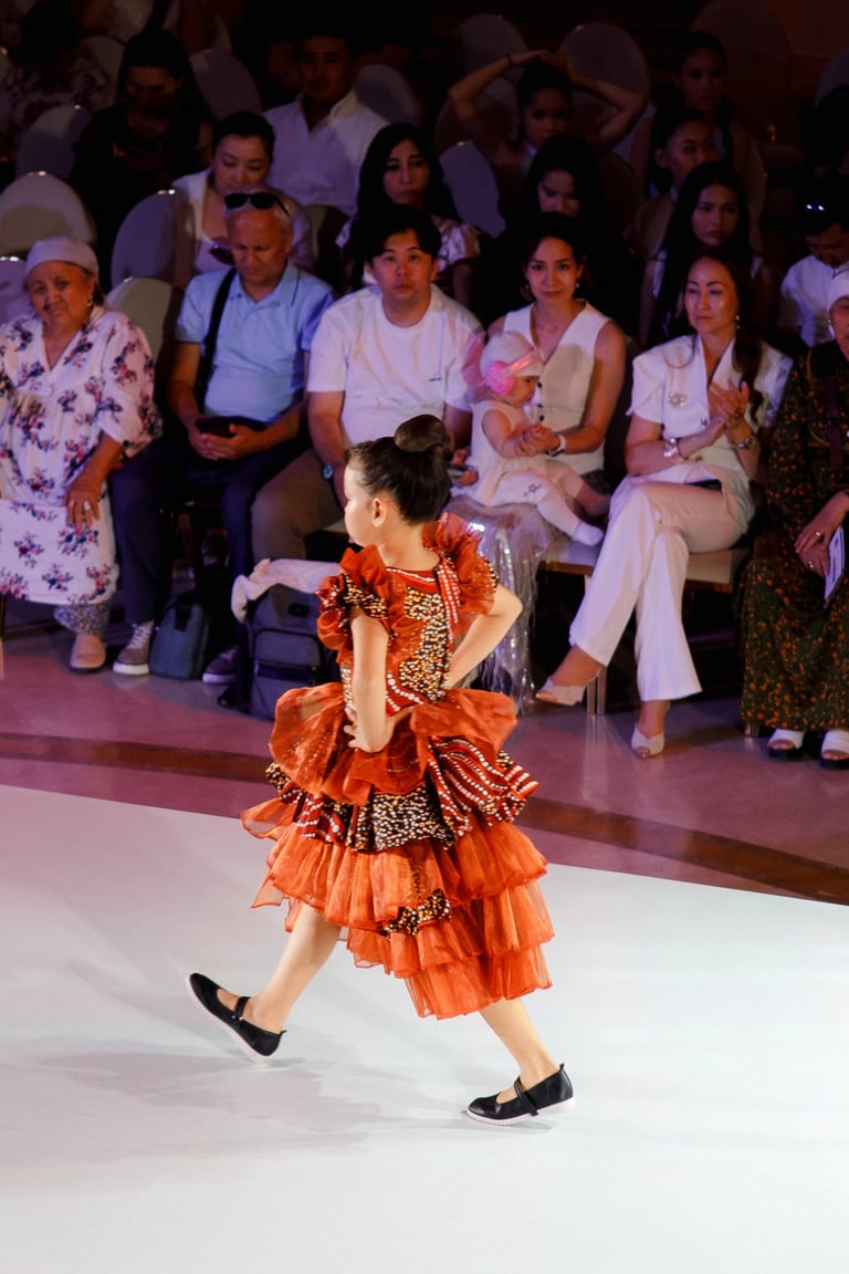 Child in orange ruffled dress performing on runway with seated audience watching in background