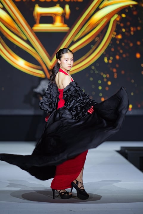 Model on runway wearing red dress with black ruffled shawl and strappy heels, striking a dynamic pose with flowing fabric against golden geometric backdrop