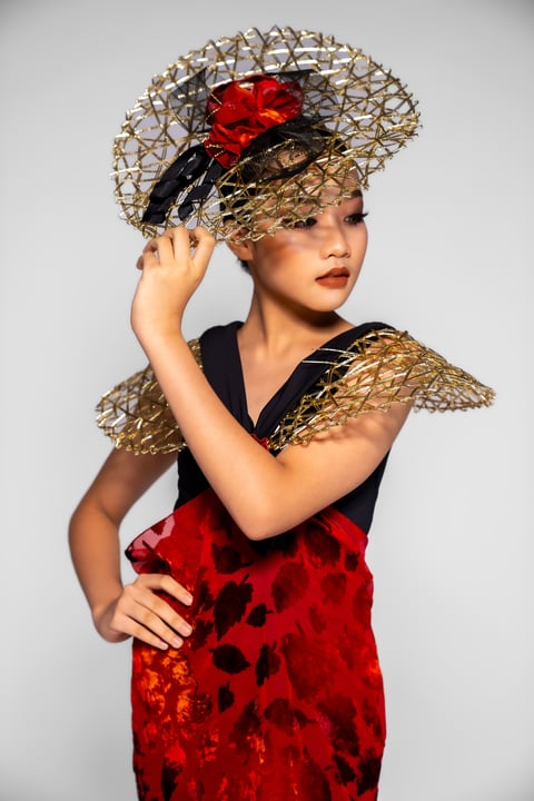 Woman in red dress with gold mesh wings and elaborate woven hat with red bow, adjusting hat against white background