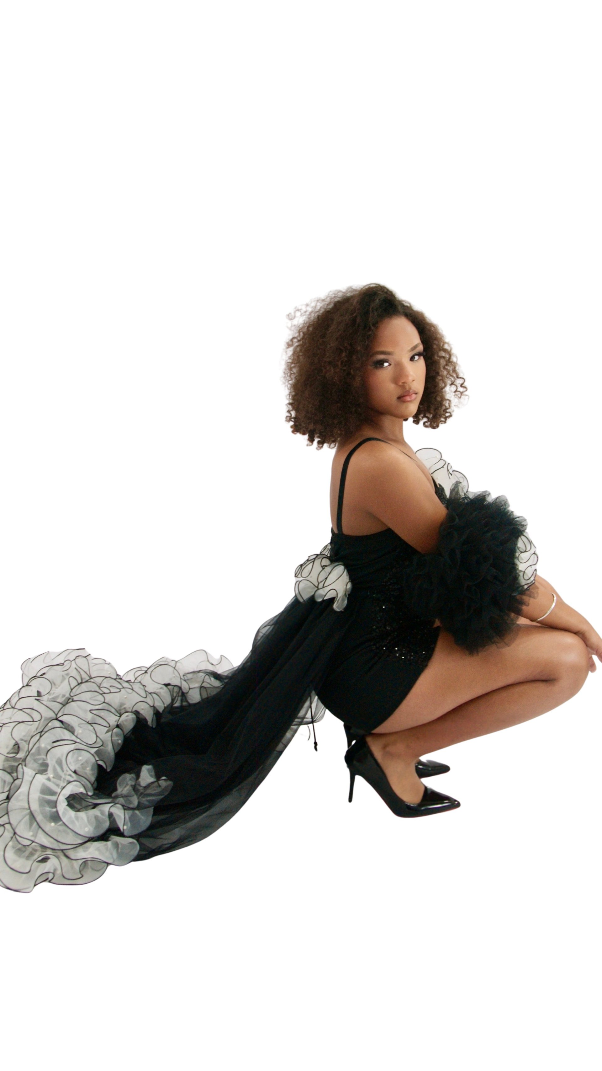 Woman with curly brown hair in elegant black tulle gown crouching against white background, looking over shoulder